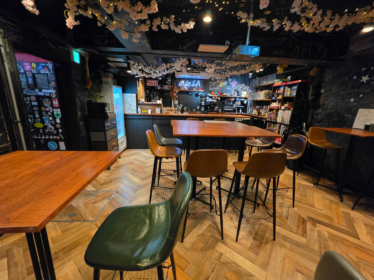 A cozy bar and cafe interior with wooden herringbone floors, high-top tables, tall stools, hanging cherry blossom decorations, and a dark counter in the background.